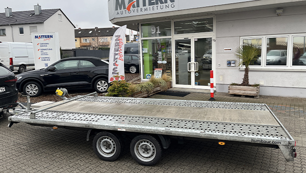 Ein leerer silberner Autotransportanhänger steht vor einem hellgrauen Gebäude