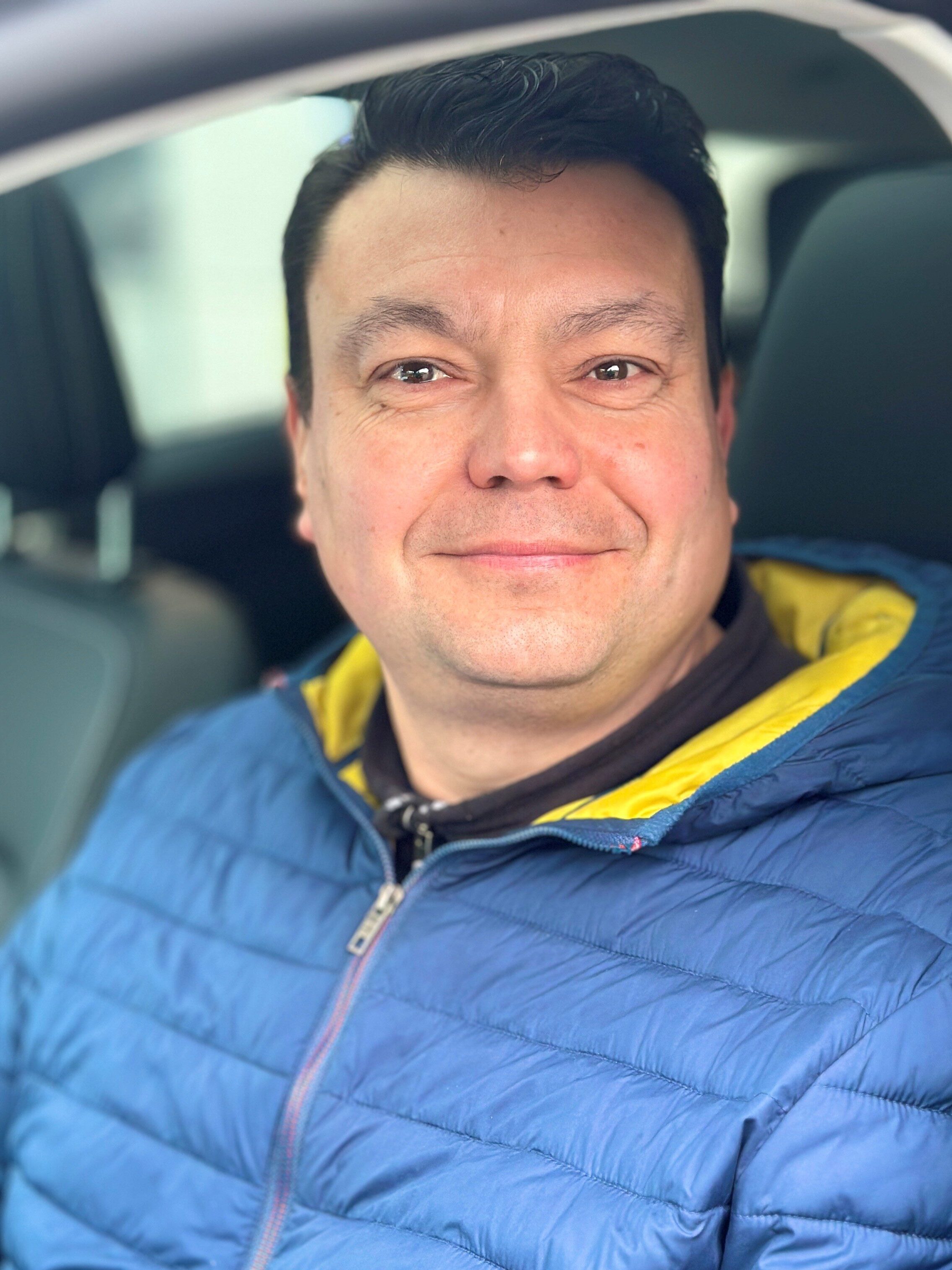Mann im Auto lächelt freundlich, trägt blaue Jacke mit gelbem Kragen, dunkles Haar.