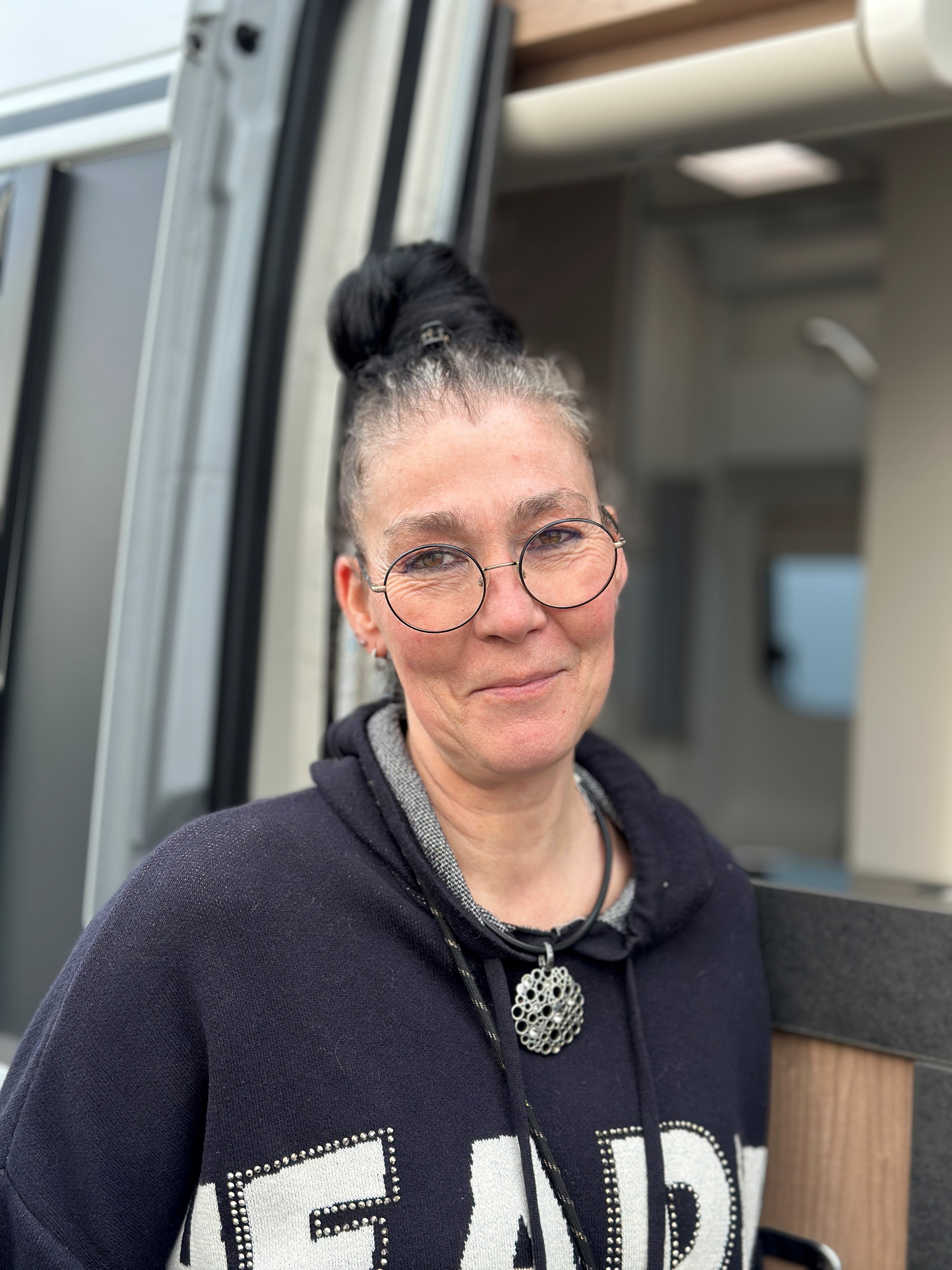 Eine Frau mit Brille lächelt freundlich. Schwarzer Pullover, hochgestecktes Haar. Wohnmobil im Hintergrund.