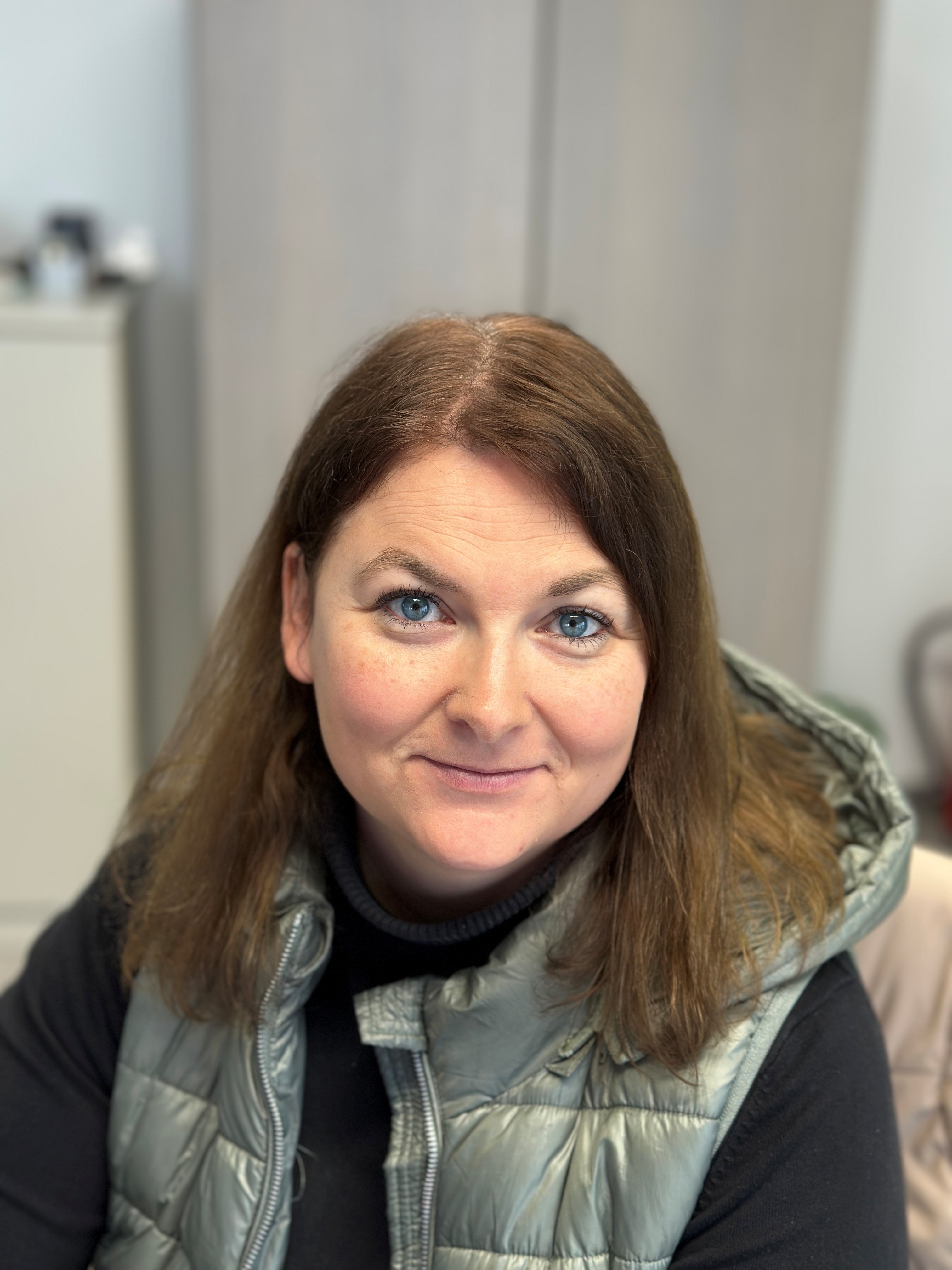 Eine Frau mit braunem Haar lächelt, trägt eine graue Weste; neutrale Hintergrundumgebung.