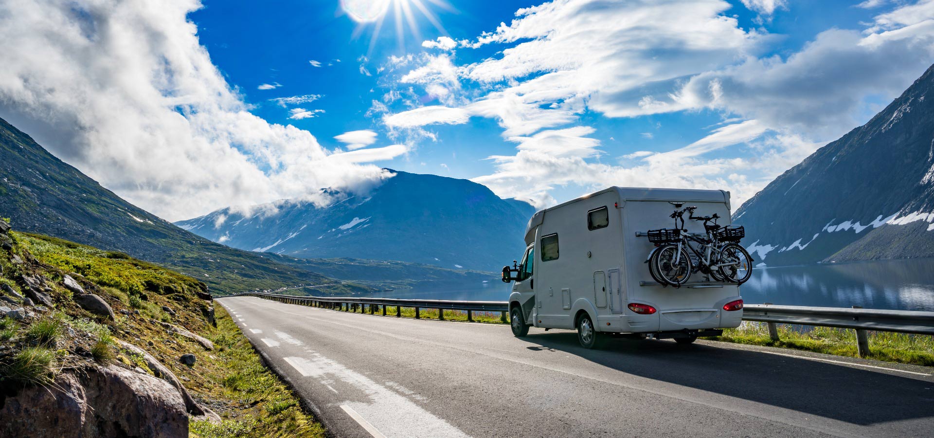 Wohnmobil auf einer malerischen Bergstraße unter sonnigem Himmel, Fahrräder auf der Rückseite.