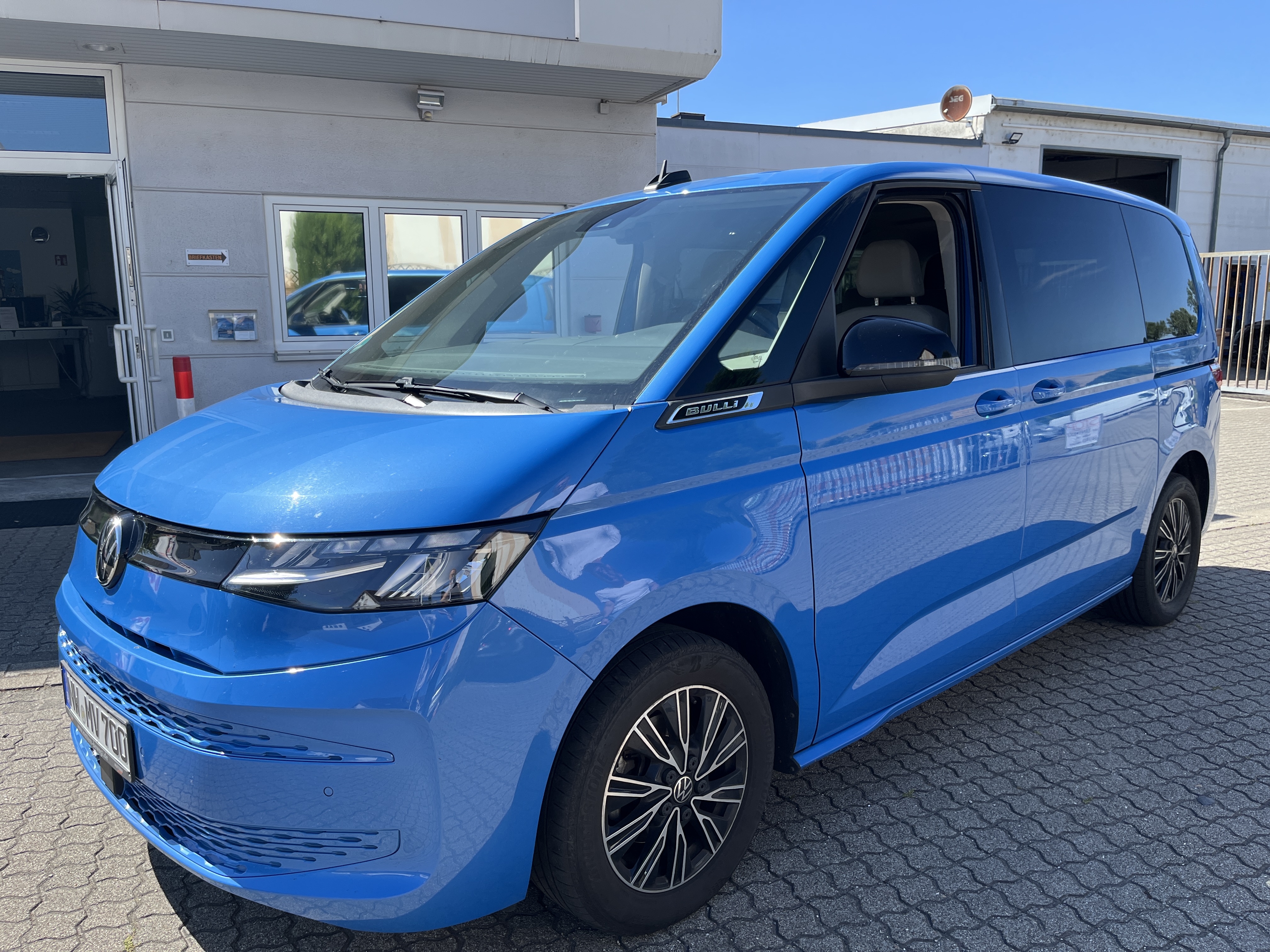 Ein blauer VW Multivan steht im Sonnenlicht vor einem modernen Industriebau