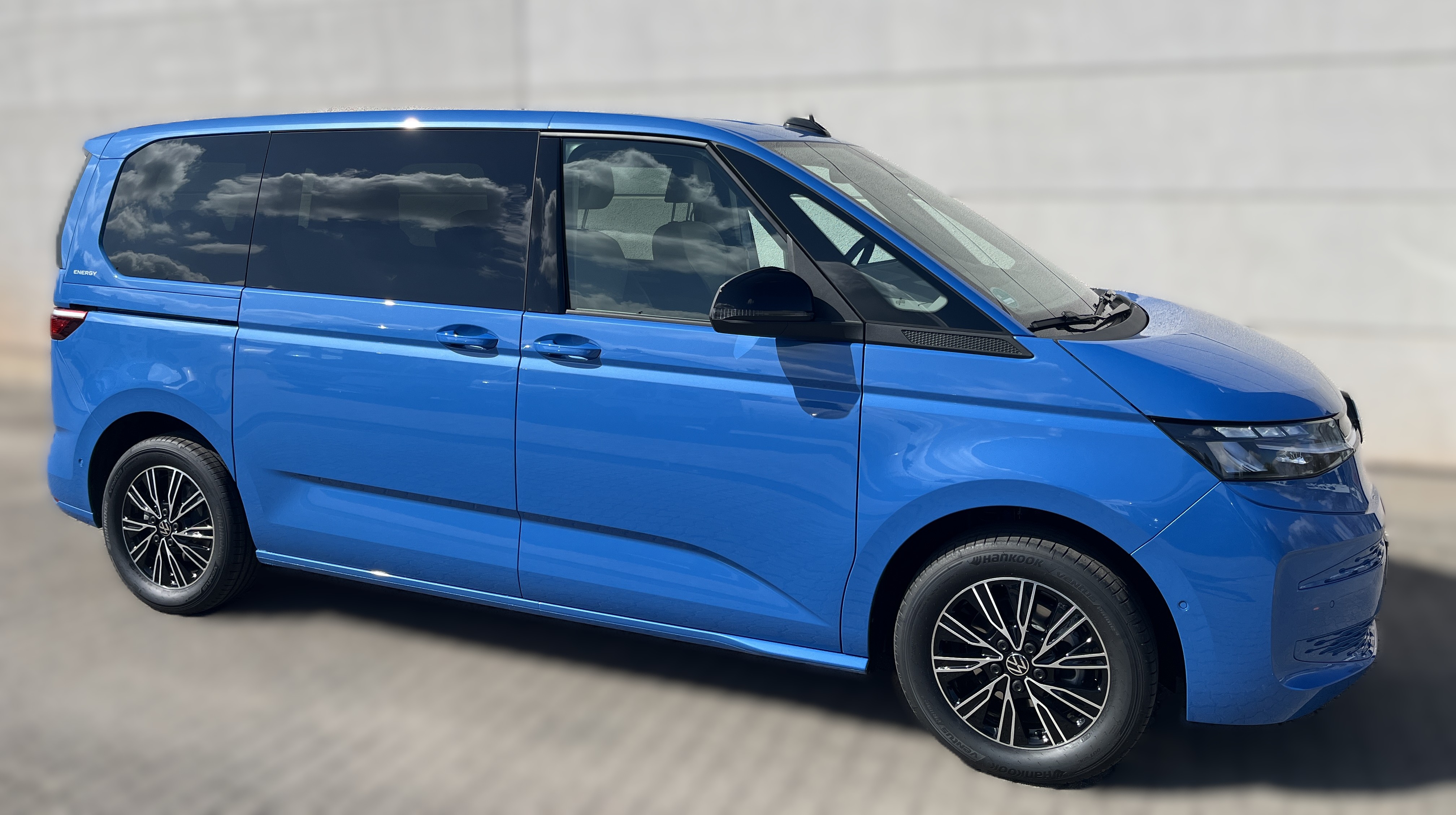 Ein blauer Van mit schwarzen Felgen steht vor einer grauen Wand im Sonnenlicht