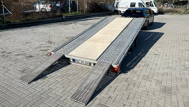 Ein silberner Autoanhänger mit Rampen steht offen auf einem gepflasterten Platz