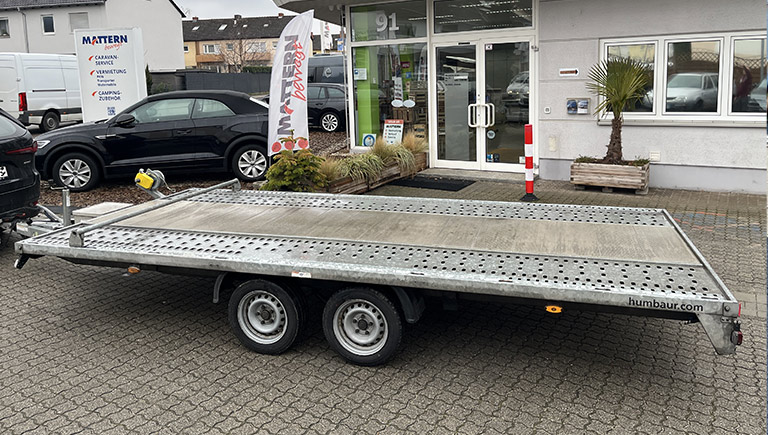 Flacher silberner Autotransportanhänger mit zwei Achsen steht vor einem Autohaus