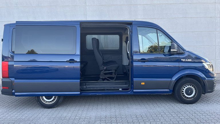 Ein blauer Transporter mit geöffneter Seitentür steht auf grauem Pflaster vor einer Wand